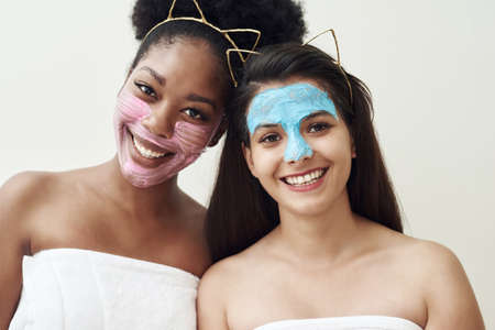 Two womans of different nationalities in cosmetic moisturizing and clay masks stand side by side and smile. Spa skin care concept.の写真素材