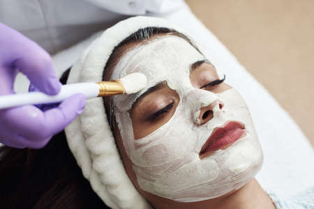 An excellent beautician applies a white anti-aging moisturizing mask to the face with a brush.の写真素材