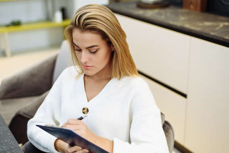 A lovely young woman is sitting at the table in her kitchen and writing in a notebook. Making a home shopping list.の写真素材