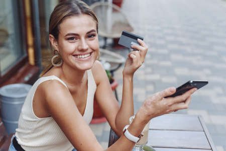 Smiling woman holding phone and credit card outdoors in cafe. A beautiful model looks at the camera and pays for purchases with a card and phone.の写真素材