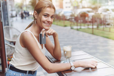 Smiling woman holding phone and credit card outdoors in cafe. A beautiful model looks at the camera and pays for purchases with a card and phone.の写真素材