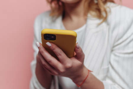 A woman holds a phone in a yellow case in her hands on a pink background. Write SMS and do shopping on the Internet.の写真素材