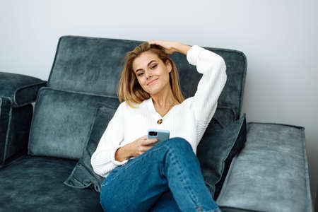 Happy young woman holding cellphone sitting on sofa and looking at camera. Female customer using tech apps on smartphone at home, enjoying easy online shoppingの写真素材