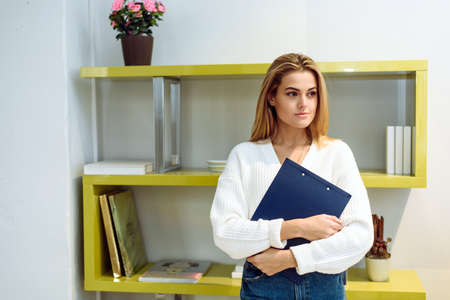 A woman holds a tablet folder for papers in her hands. A lovely lady stands in the office and looks at the cameraの写真素材