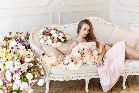 Young lady Caucasian white woman with blonde high-heeled hairstyle and bare shoulders posing with different colors in studio interiors with sofaの写真素材
