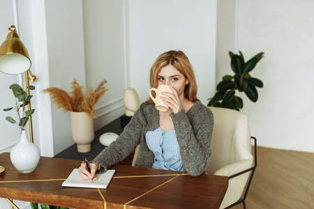Smiling blonde young woman in casual doing notes in daily book with cup of tea on kitchen at homeの写真素材