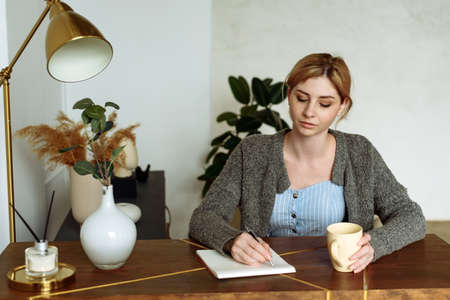 A cute young woman makes notes in a notebook while sitting at her desk at home and drinking coffeeの写真素材