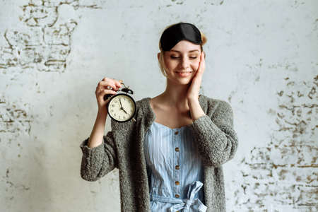 Sleepy girl in a sleep mask holds an alarm clock in her hand and yawnsの写真素材
