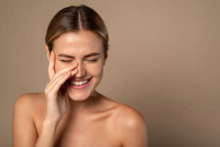 Skin care. Woman with beauty face touching healthy facial skin. Beautiful smiling Caucasian female model with natural makeup touching glowing hydrated skin on beige backgroundの写真素材