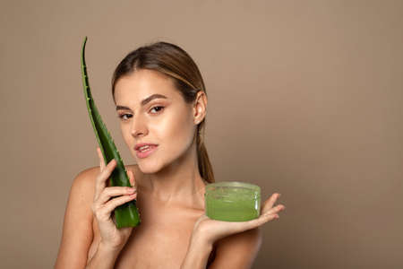 Close up beauty portrait of attractive young topless woman with brown hair isolated on beige background holding aloe leaf and gel in jar. Face skin care conceptの写真素材