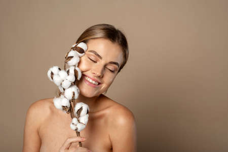 Portrait of pretty young woman with branch of soft cotton with clean fresh skin in studio against beige background. Cosmetology, beauty and spa concept.の写真素材