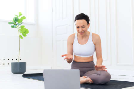 A young female trainer teaches yoga remotely over the Internet. The woman shows exercises in the gym on the mat. The concept of distance learningの写真素材