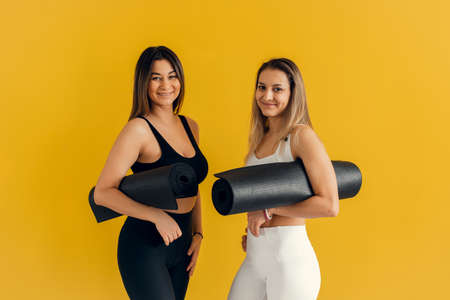 Two athletic women in sportswear hold rugs close at hand and smile at the camera on a yellow background. Sports and recreation conceptの写真素材