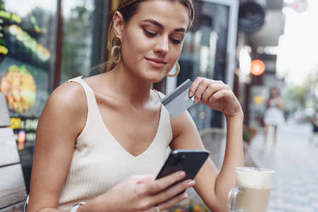 Woman sitting at cafe holding mobile phone and plastic card signing up on website.Beautiful model paying with credit card while shopping online using mobileの写真素材