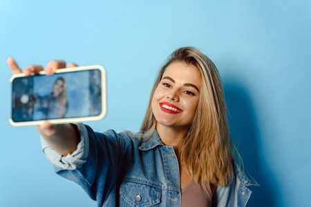 Portrait of a happy young girl with glitter on her cheeks taking a selfie, isolated on blue background. Image of excited happy young woman taking selfie with cameraの写真素材