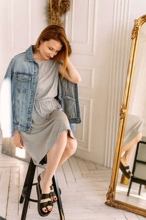 Cozy home atmosphere. Stylish redhead woman in gray dress and jeans sits on a chair at home next to a mirror and wheat. Portrait beautiful young woman happy smile relax in living room interiorの写真素材