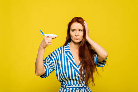 Young beautiful redhead woman holding airplane over yellow background with angry face, negative sign, rejection concept. The woman is angry from lost luggage, an unsuccessful vacation or flightの写真素材