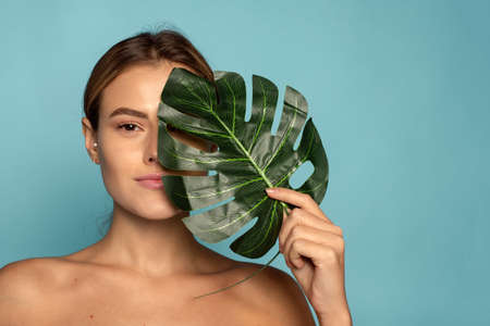 A lovely female model covers part of her face with a tropical palm leaf. Skin care, hydration. Cosmetics with natural ingredientsの写真素材