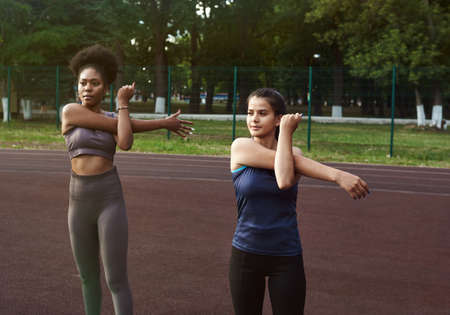Two women of different nationalities warm up before training outdoors. Warming up muscles before strength training or joggingの写真素材