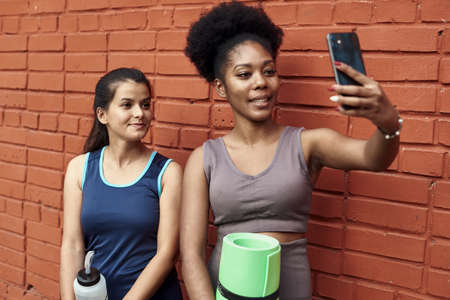 Image of amazing young athletic women taking a selfie against a brick wall. Smiling black women spend time together after sports workoutの写真素材