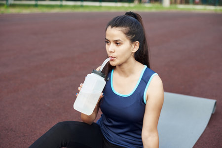 Young beautiful woman sits on a yoga mat outdoors and drinks water from a reusable water bottle. Outdoor sportsの写真素材