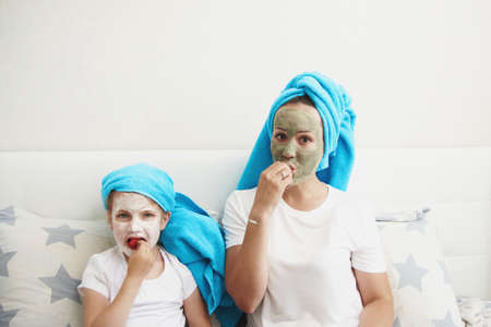 Morning care routine. Mom and daughter conduct beauty treatments. They are sitting on the couch and applying pieces of cucumber to their eyes.の写真素材