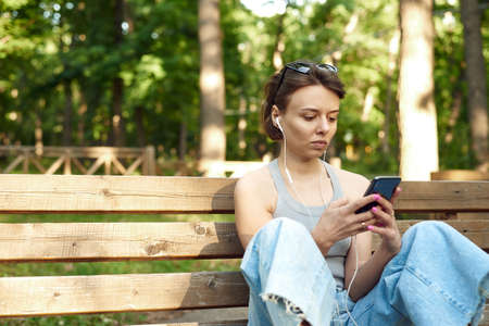 Cheerful millennial blonde teenage girl, sitting on bench outdoors, with smart phone and headphones. Young female student in park smiling and relaxingの写真素材