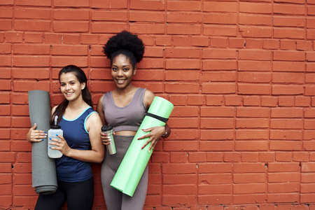 Two lovely young women in tracksuits stand against a brick wall in the street. The girlfriends are smiling while holding yoga mats and water bottlesの写真素材