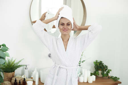 Lovely happy young woman in a bathrobe straightens a white towel turban on her head. Spa concept, skin care and self-loveの写真素材