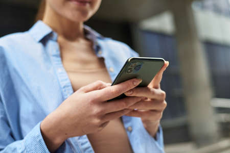 Female manager uses mobile phone in city outdoors, women in blue shirt and jeans writes message on cellphone. Happy business woman chatting on phoneの写真素材