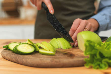 A woman cuts cucumbers and apples in the kitchen for breakfast. A young housewife stands in her kitchen. Close upの写真素材