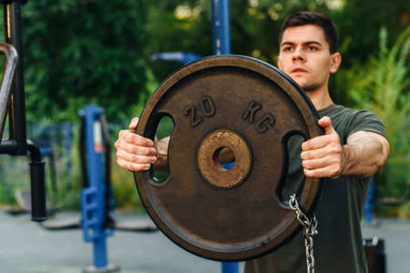 A young man goes in for sports, pumps muscles with the help of a heavy barbell disc. Outdoor. Sports conceptの写真素材