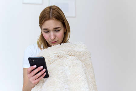 Portrait of a beautiful young woman with a depressed expression, sitting on the bed, holding her phone. Cyber bullying, smartphone addiction and depression, breakup, sad newsの写真素材