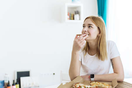 Portrait of a beautiful woman eating pizza in the kitchen. Wrong food, fast food conceptの写真素材