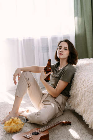 A beautiful woman switches channels and films on a TV using a remote control. She drinks beer and eats chips at home. Relax at home watching TV and moviesの写真素材