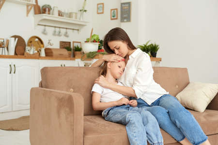 A young mother supports her daughter, calms her down and strokes her head. Little girl crying and complaining to her mother on the couch at homeの写真素材