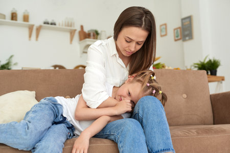 A young mother supports her daughter, calms her down and strokes her head. Little girl crying and complaining to her mother on the couch at homeの写真素材