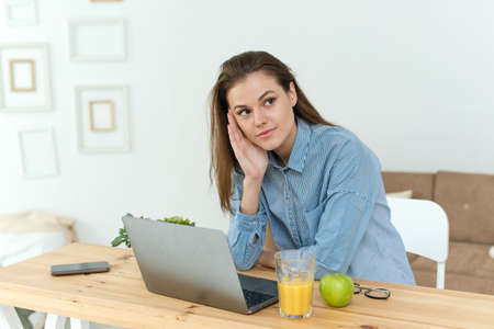 Student thinking while doing complicated homework on line with a laptop at home. Photo of young pretty businesswoman sit indoors in office using laptop computer staring thoughtfully into the airの写真素材