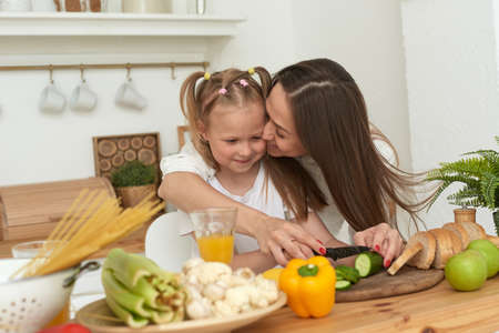 Cheerful mother and little daughter are preparing salad together in the kitchen and having fun. The girl kisses her mother at homeの写真素材