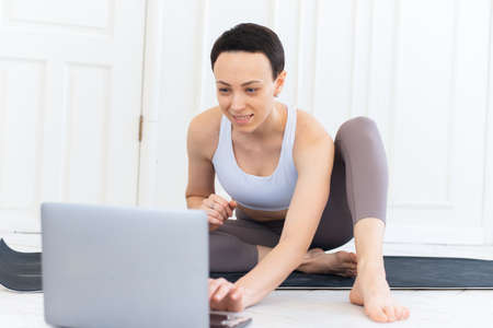 A sporty woman in sportswear is sitting on the floor and is using a laptop at home in the living room. Sport and recreation conceptの写真素材