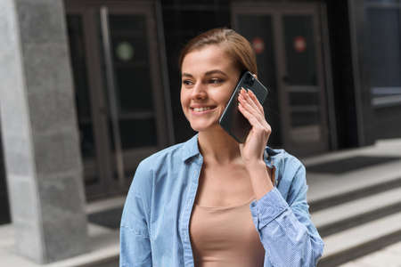 A businesswoman walks against the backdrop of an office center and speaks on the phone. Communication with business partners at a distanceの写真素材