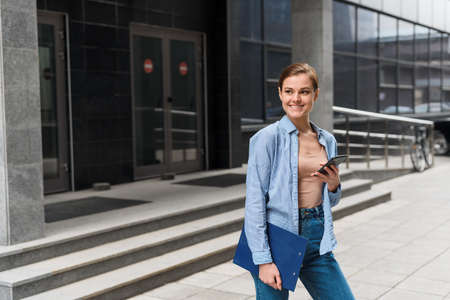 A young stylish businesswoman walks against the background of the business center and holds a folder with documents under her arm, a phone in her hand. Going to work after the coronavirus pandemicの写真素材