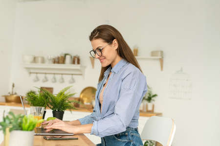 Attractive businesswoman studying online, using laptop software, web surfing information or shopping in internet store. Smiling young woman using laptop in the kitchen at home. Working from home in quarantine lockdown.の写真素材