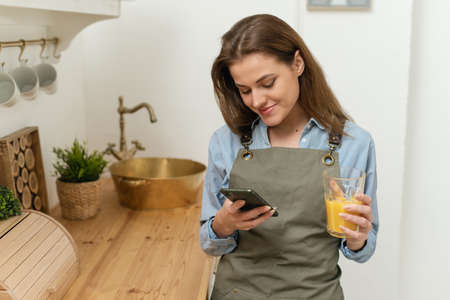 Portrait of cheerful young woman standing in kitchen with cup of fresh orange juice and checking text messages on her phoneの写真素材