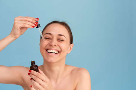 Smiling woman holding vitamin c serum near her face on blue background. Skin care and health conceptの写真素材
