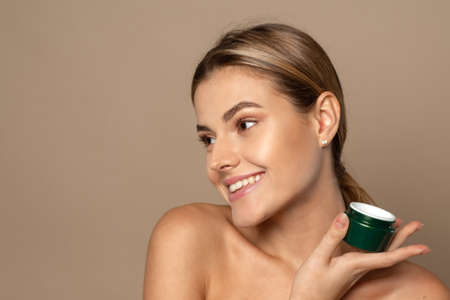 Cute woman holding a jar of moisturizer in the studio. Skin care and hydration concept. Portrait of beautiful woman applying some cream to her face for skin careの写真素材