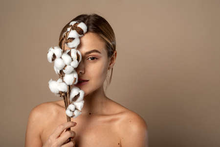 Portrait of pretty young woman with branch of soft cotton with clean fresh skin in studio against beige background. Cosmetology, beauty and spa concept.の写真素材