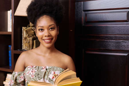 Portrait of a young black female student. She is standing in the university library, holding a book and looking at the cameraの写真素材