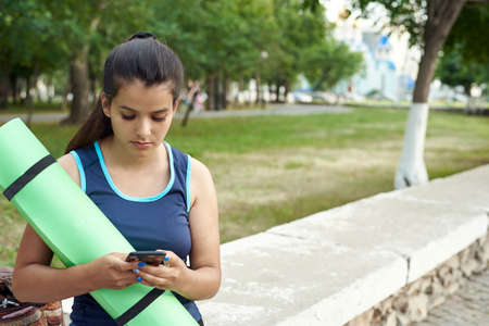A young woman holds a yoga mat and climbs on the phone after a workout on the streetsの写真素材
