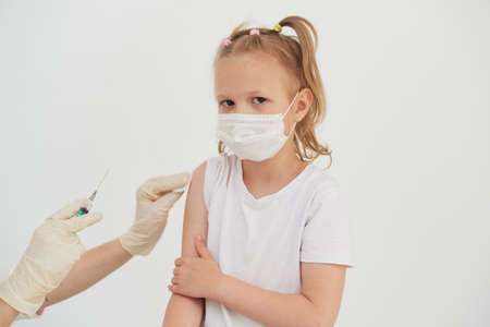 Vaccination concept. Doctors hand vaccinates a cute little girl in a protective mask on a white background, close-upの写真素材